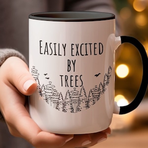 May include: White ceramic mug with a black handle. The mug has a black rim and a black line around the top. The mug has a black and white design of trees and the text "Easily Excited By Trees".