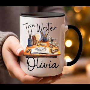 May include: White ceramic mug with a black interior and handle. The mug features the text "The Writer Olivia" in black script. A watercolor illustration of a writer's desk with books, pens, and glasses is also on the mug.