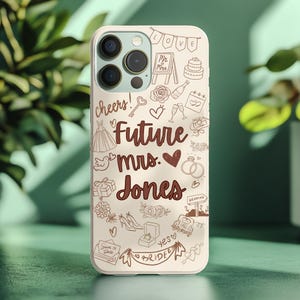 May include: A beige phone case with a wedding theme. The case features the words "Future mrs. Jones" in a dark brown script, along with wedding-related doodles such as a cake, rings, and a dress. The word "cheers!" is also visible.