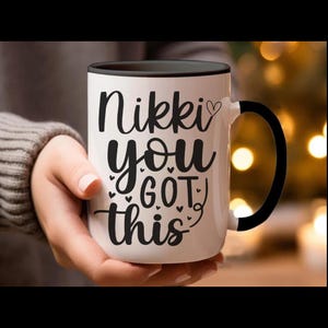 May include: White ceramic mug with a black interior and handle. The mug features the text "Nikki, you got this" in black script with heart and dot accents. The mug is held by a person wearing a gray sweater.