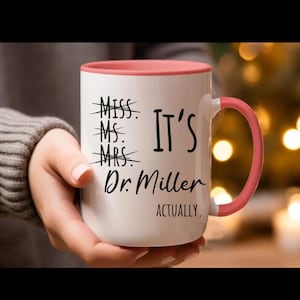 May include: White ceramic mug with a pink rim and handle. The mug has black text that reads "Miss, Ms, Mrs. It's Dr. Miller Actually."