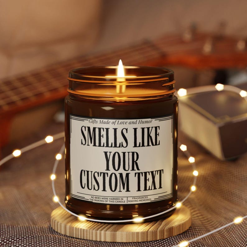 May include: A lit brown glass jar candle with the text "SMELLS LIKE YOUR CUSTOM TEXT" on a white label. The candle sits on a wooden coaster, surrounded by string lights. The candle's label also includes the text "Gifts Made of Love and Humor".