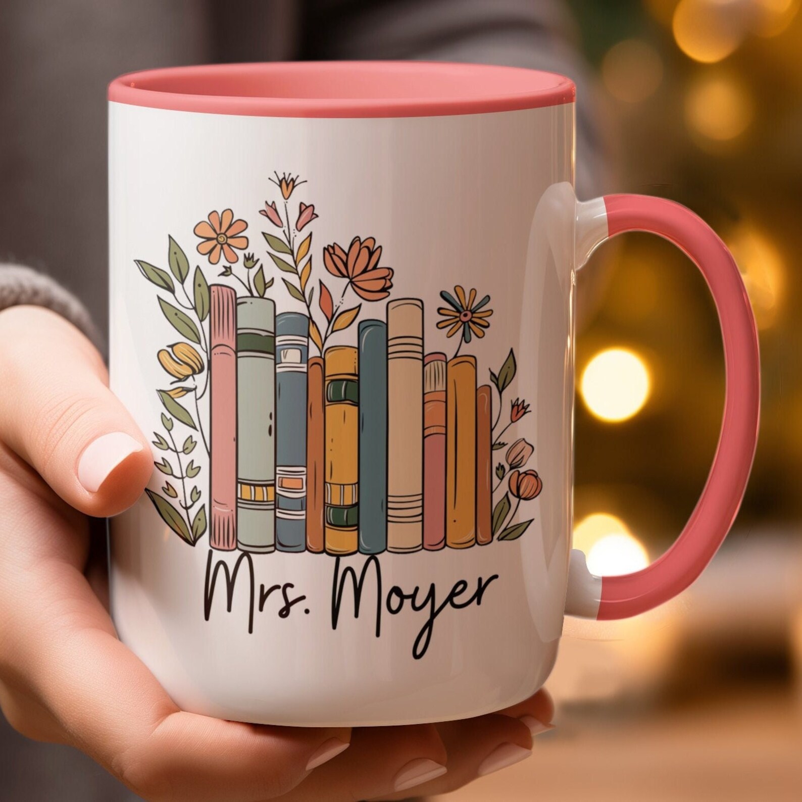 A white ceramic mug with a coral pink interior and matching handle is shown held in both hands. The design on the mug features a colorful row of illustrated books with pastel-colored spines in shades of teal, mustard yellow, dusty rose, and soft orange. The books are surrounded by whimsical hand-drawn flowers and leafy vines in coordinating tones, adding a playful botanical flair. Beneath the bookshelf illustration, the name “Mrs. Moyer” is printed in a bold, flowing black script.