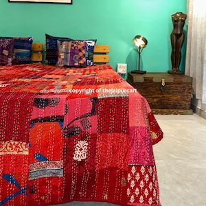 May include: A red patchwork quilt with various patterns and colors covers a bed. The quilt is the focal point, with a headboard and pillows visible. A wooden chest and a statue are in the background.