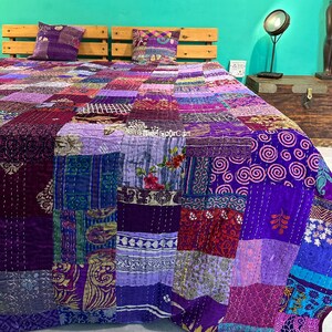 Peut inclure: Une courtepointe patchwork aux tons de violet, rose et bordeaux recouvre un lit. La courtepointe est composée de divers carrés de tissu aux motifs et textures variés. Deux coussins violets assortis reposent contre une tête de lit en bois.