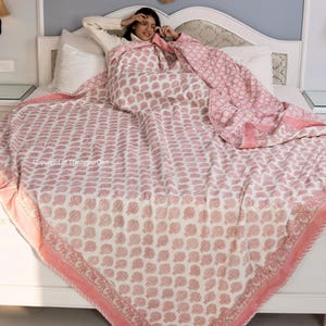 May include: A pink and white cotton quilt with a floral print. The quilt is folded on a bed with a white headboard.