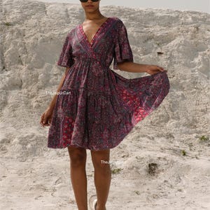 K&ouml;nnte beinhalten: Kurzes, gemustertes Kleid in Burgunder-, Rosa- und Lilat&ouml;nen. Das Kleid hat einen V-Ausschnitt, kurze &Auml;rmel und einen Stufenrock. Die Person tr&auml;gt eine Sonnenbrille und wei&szlig;e Turnschuhe. The Jaipur Cart ist auf dem Kleid aufgedruckt.
