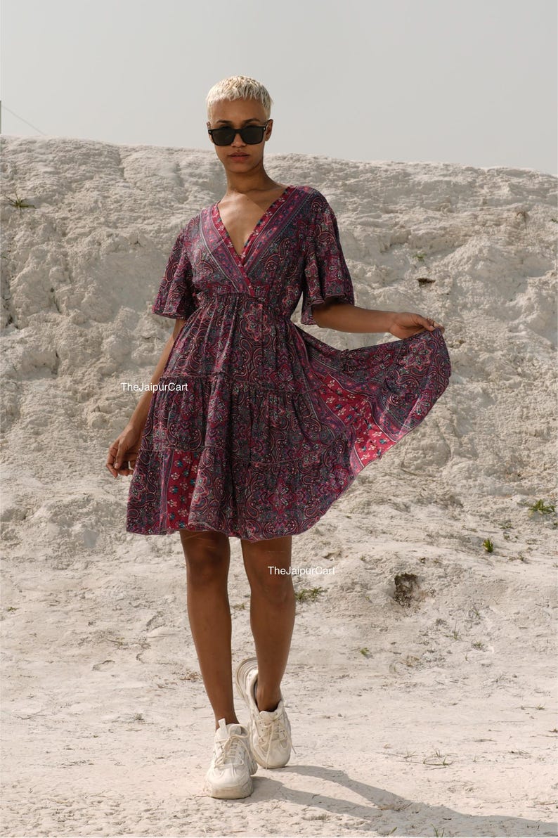 K&ouml;nnte beinhalten: Kurzes, gemustertes Kleid in Burgunder-, Rosa- und Lilat&ouml;nen. Das Kleid hat einen V-Ausschnitt, kurze &Auml;rmel und einen Stufenrock. Die Person tr&auml;gt eine Sonnenbrille und wei&szlig;e Turnschuhe. The Jaipur Cart ist auf dem Kleid aufgedruckt.