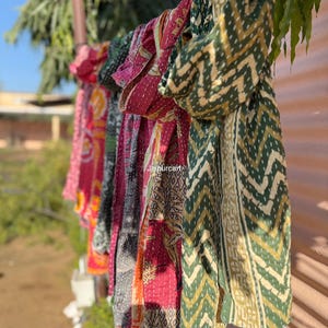 Puede incluir: Una fila de coloridas bufandas colgadas al aire libre. Las bufandas presentan varios patrones y colores, incluyendo rojo, verde y amarillo. Algunas tienen diseños geométricos, mientras que otras tienen patrones florales o abstractos. Las bufandas están hechas de una tela ligera.