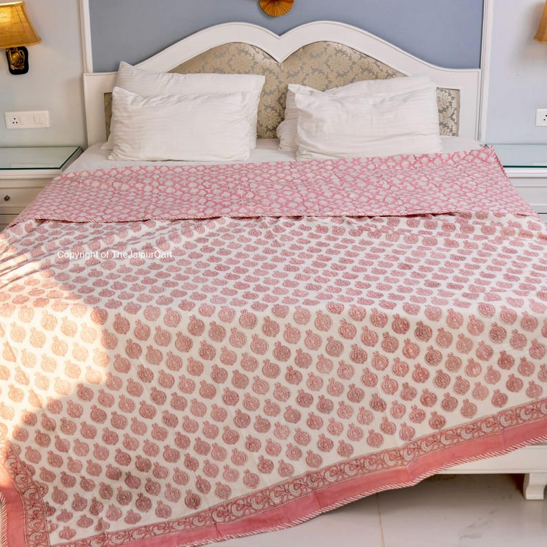 May include: A white cotton quilt with a pink floral pattern. The quilt is folded on a bed with white pillows.