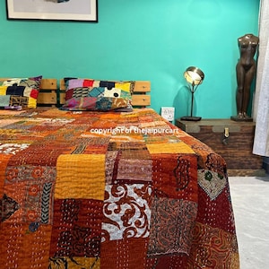 May include: A colorful patchwork quilt with a variety of patterns and colors, including orange, brown, and white. The quilt is made of recycled saris and is hand-stitched. It is laid out on a bed with pillows and a wooden headboard.