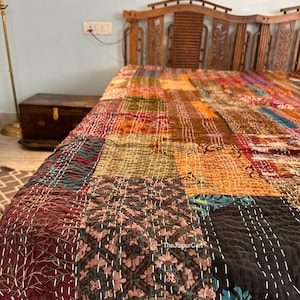 Peut inclure: Une courtepointe patchwork avec une variété de couleurs et de motifs, notamment le rouge, le brun, l'or et le bleu. La courtepointe est faite de différents tissus et est cousue avec du fil blanc. La courtepointe est sur un cadre de lit en bois.