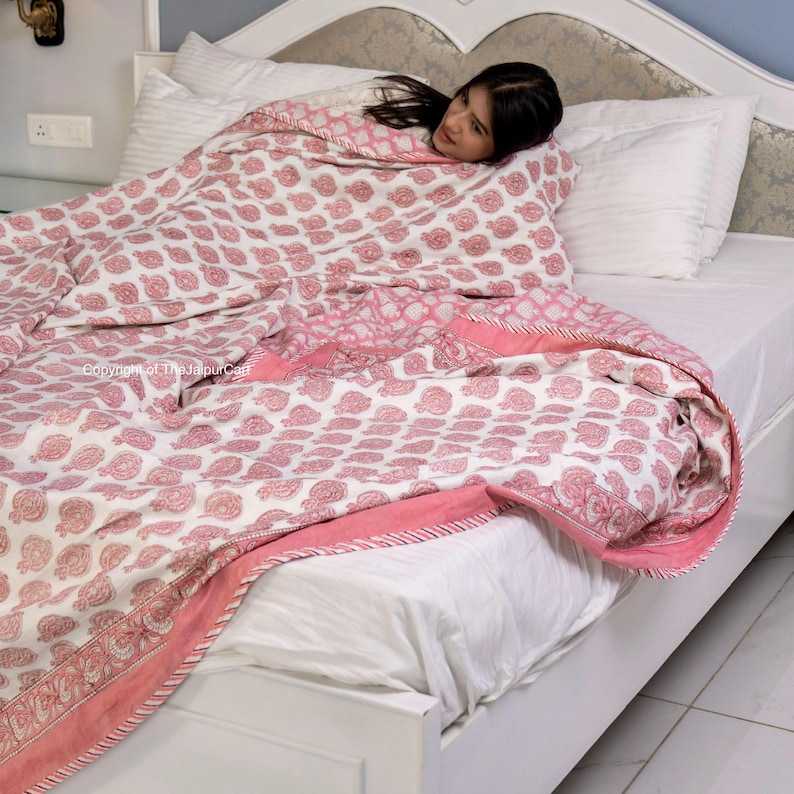 May include: A pink and white cotton quilt with a floral pattern. The quilt is folded on a white bed with a white pillow.