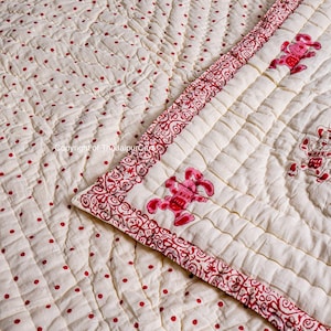 K&ouml;nnte beinhalten: Eine wei&szlig;e Baumwolldecke mit rotem Tupfenmuster und rotem Blumenrand. Die Decke ist in einem Rautenmuster gesteppt und zeigt rote gestickte Tiere.