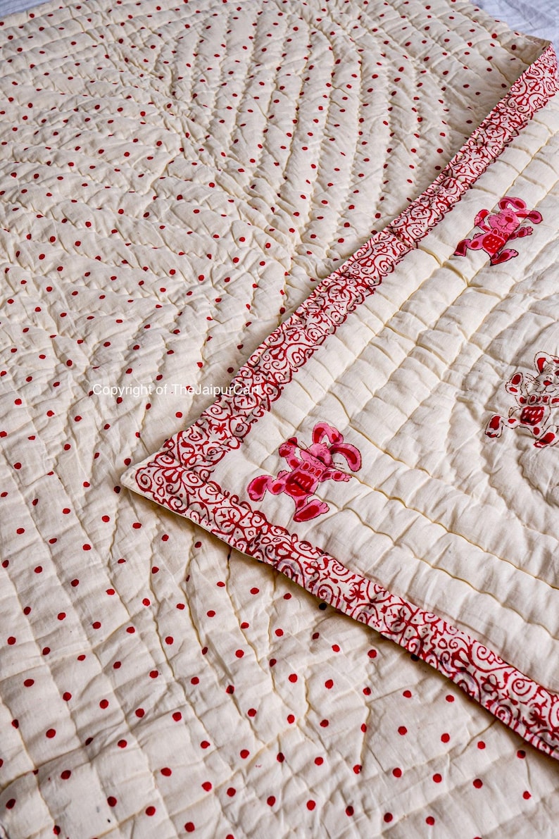 K&ouml;nnte beinhalten: Eine wei&szlig;e Baumwolldecke mit rotem Tupfenmuster und rotem Blumenrand. Die Decke ist in einem Rautenmuster gesteppt und zeigt rote gestickte Tiere.