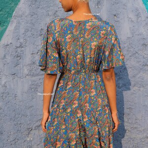 K&ouml;nnte beinhalten: Blaues Paisley-Kleid mit kurzen &Auml;rmeln und einem Stufenrock. Das Kleid hat einen V-Ausschnitt und eine betonte Taille. Der Stoff hat ein Blumen- und Paisley-Muster in Rosa-, Gelb- und Blaut&ouml;nen. The Jaipur Cart ist auf dem Kleid aufgedruckt.