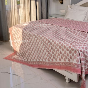 May include: A pink and white cotton quilt with a paisley print. The quilt is folded on a bed with white sheets.