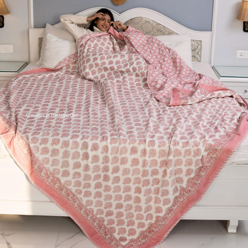 May include: A pink and white cotton quilt with a floral print. The quilt is folded on a bed with a white headboard.