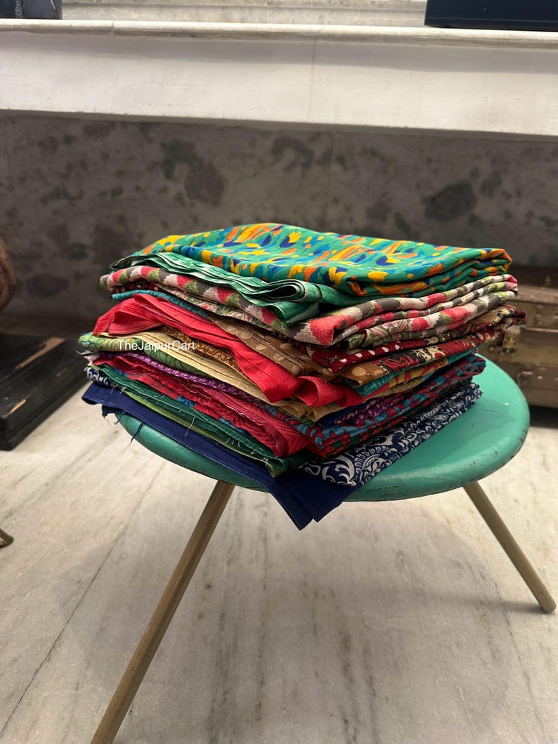 May include: A stack of colorful fabric scraps with various patterns, including floral, geometric, and paisley designs. The fabric is arranged on a teal stool with wooden legs. The text "The Jaipur Cart" is visible on the fabric.