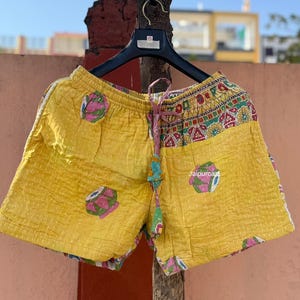 Può includere: Pantaloncini gialli trapuntati con vita con coulisse e accenti colorati. I pantaloncini presentano un motivo ripetuto di motivi floreali rosa e verdi. Il cordoncino è un mix di rosa e verde. I pantaloncini sono appesi a una gruccia nera.