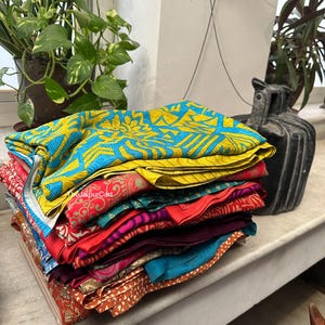 May include: A stack of colorful fabric scraps in various patterns and colors, including yellow, blue, red, pink, and orange. The fabric is folded neatly and arranged in a pile on a white surface.