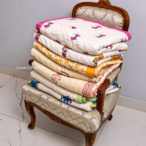 K&ouml;nnte beinhalten: Ein Stapel von sechs bunten Baumwolldecken mit verschiedenen Mustern, darunter Sterne, Tiere und Blumenmuster, wird auf einem Vintage-Stuhl mit hellgrauer Stoffpolsterung pr&auml;sentiert.