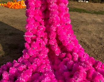 May include: A cascade of vibrant pink floral garlands, densely packed with small, fluffy blooms. The garlands are arranged in a vertical display, with green stems visible. Other garlands in red, yellow, and orange are visible in the background.