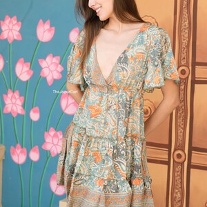 K&ouml;nnte beinhalten: Gemustertes Kleid mit kurzen &Auml;rmeln, V-Ausschnitt und Stufenrock. Das Kleid hat ein Blumenmuster in Blau-, Orange- und Graut&ouml;nen. Der Text "The Jaipur Cart" ist auf dem Kleid zu sehen. Der Hintergrund zeigt eine blaue Wand mit rosa Lotusblumen.