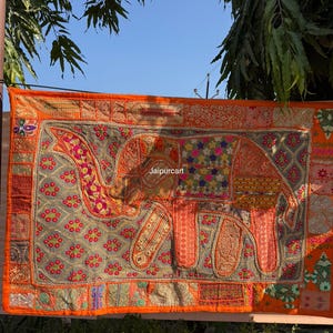 May include: A vibrant orange and multi-colored textile wall hanging featuring an embroidered elephant design. The elephant is detailed with various patterns and colors, including pink, green, and gold. The text "Jaipurcart" is visible.