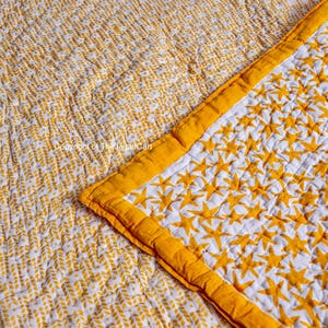 K&ouml;nnte beinhalten: Eine wei&szlig;e und gelbe Baumwolldecke mit einem Sternenmuster auf der einen Seite und einem Blattmuster auf der anderen Seite. Die Decke ist in der Mitte gefaltet und zeigt beide Seiten.