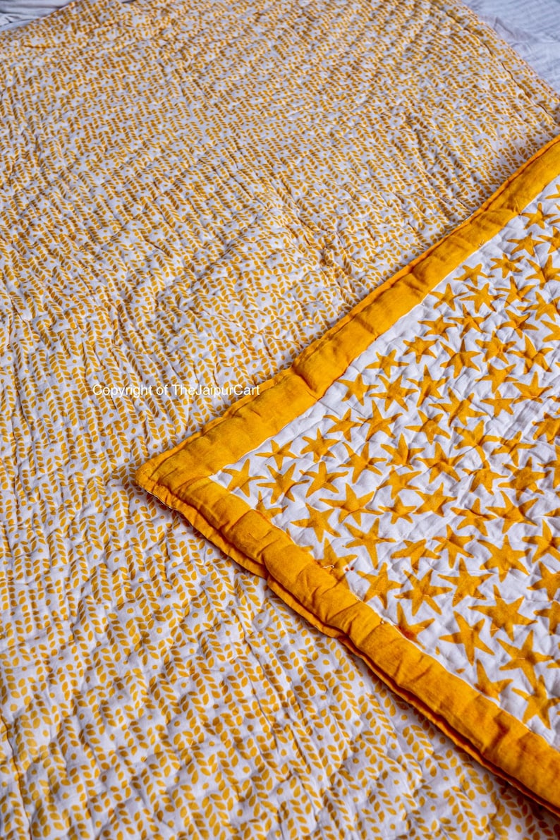 K&ouml;nnte beinhalten: Eine wei&szlig;e und gelbe Baumwolldecke mit einem Sternenmuster auf der einen Seite und einem Blattmuster auf der anderen Seite. Die Decke ist in der Mitte gefaltet und zeigt beide Seiten.
