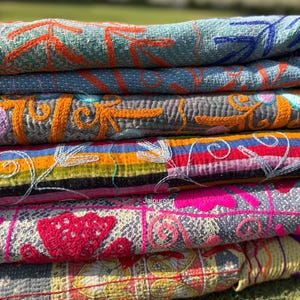 Può includere: Una pila di tessuti ricamati colorati. I tessuti presentano motivi intricati in tonalità di blu, arancione, rosso, rosa e giallo. I tessuti sono impilati uno sull'altro, con i bordi di ogni pezzo visibili.