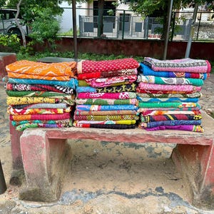 Pode incluir: Uma pilha de colchas kantha coloridas, dobradas e dispostas em um banco de pedra. As colchas são feitas de vários tecidos e padrões, incluindo desenhos florais, geométricos e paisley.