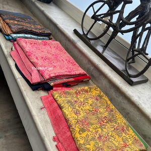 May include: A stack of colorful fabric scarves with floral patterns. The scarves are folded and arranged on a light gray surface. The scarves are in shades of pink, blue, yellow, and brown.