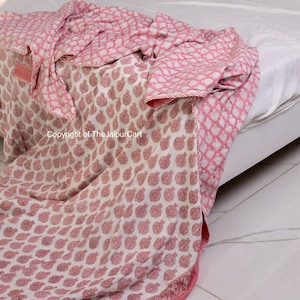 May include: A pink and white cotton quilt with a floral print. The quilt is folded on a bed and has a border of pink and white.