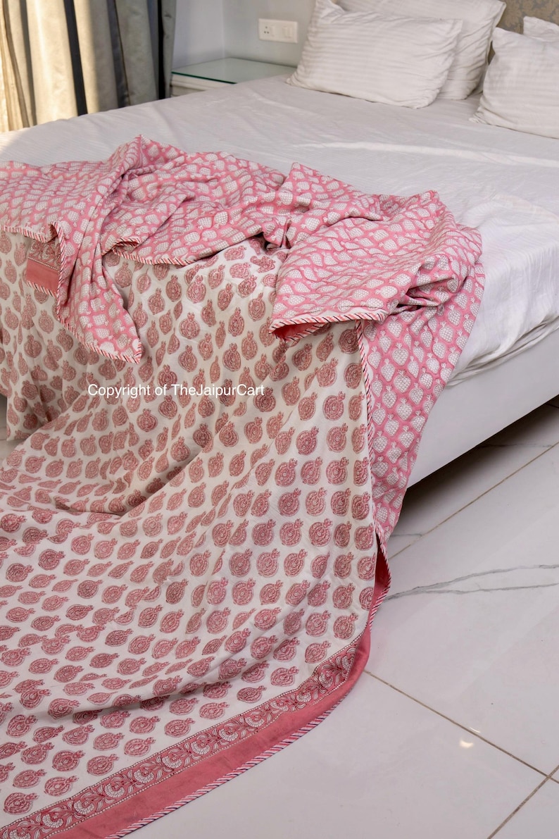 May include: A pink and white cotton quilt with a floral print. The quilt is folded on a bed and has a border of pink and white.