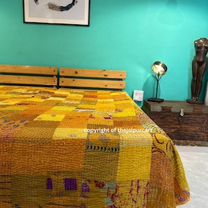 May include: A patchwork quilt in shades of yellow and gold covers a bed. The quilt features a variety of stitched squares and rectangles. A wooden headboard is visible. The wall behind the bed is teal, and a framed artwork hangs above the headboard.