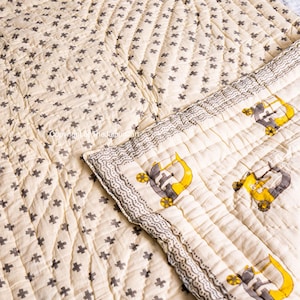 K&ouml;nnte beinhalten: Eine cremefarbene Steppdecke mit einem grauen Kreuzmuster. Die Steppdecke ist in der Mitte gefaltet und zeigt ein cremefarbenes Stoff mit einem grauen und gelben LKW-Muster.