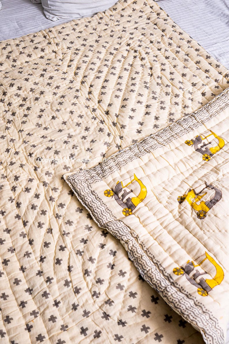K&ouml;nnte beinhalten: Eine cremefarbene Steppdecke mit einem grauen Kreuzmuster. Die Steppdecke ist in der Mitte gefaltet und zeigt ein cremefarbenes Stoff mit einem grauen und gelben LKW-Muster.