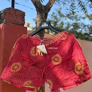 May include: Red shorts with a drawstring waist and a decorative pattern of yellow circles. The shorts are hanging on a black hanger against a blurred outdoor background. The text "Jaipurcart" is visible on the shorts.
