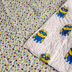 K&ouml;nnte beinhalten: Eine wei&szlig;e Babydecke aus Baumwolle mit einem bunten Blattmuster auf der einen Seite und einer wei&szlig;en Seite mit einem sich wiederholenden Muster von gelben Minions, die blaue Schutzbrillen tragen.