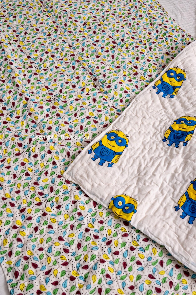 K&ouml;nnte beinhalten: Eine wei&szlig;e Babydecke aus Baumwolle mit einem bunten Blattmuster auf der einen Seite und einer wei&szlig;en Seite mit einem sich wiederholenden Muster von gelben Minions, die blaue Schutzbrillen tragen.