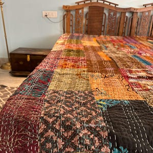 May include: A colourful patchwork quilt featuring a variety of patterns and colours, including floral and geometric designs. The quilt is made from different fabric squares stitched together with white thread. The quilt is on a wooden bed frame.