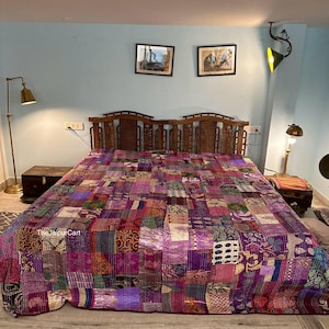 May include: A patchwork quilt in shades of purple, pink, and red covers a bed. The quilt is made of various fabric squares, creating a textured and colourful design. The headboard is made of wood. The room has a light blue wall.