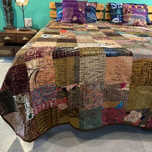 Puede incluir: Una colcha de retazos hecha de una variedad de telas en tonos marrones, beige y morados. La colcha est&aacute; colocada sobre una cama con dos almohadas decorativas.