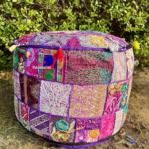 Peut inclure: Un pouf rond en patchwork dans les tons de violet, rose et or. Le pouf est composé de divers carrés de tissu avec des détails brodés et texturés. The Jaipur Cart est imprimé sur le côté. Des pompons ornent les bords.