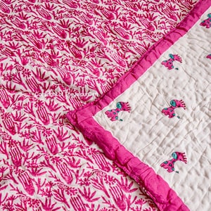 K&ouml;nnte beinhalten: Eine rosafarbene und wei&szlig;e Baumwolldecke mit einem handbedruckten Muster aus rosa Blumen und Bl&auml;ttern. Die Decke hat einen rosa Rand und einen wei&szlig;en Hintergrund mit kleinen gestickten Giraffen.