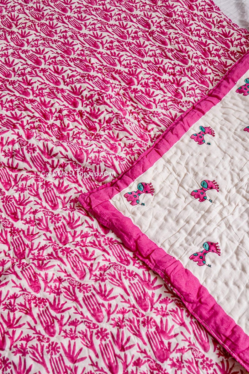 K&ouml;nnte beinhalten: Eine rosafarbene und wei&szlig;e Baumwolldecke mit einem handbedruckten Muster aus rosa Blumen und Bl&auml;ttern. Die Decke hat einen rosa Rand und einen wei&szlig;en Hintergrund mit kleinen gestickten Giraffen.