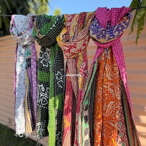 Puede incluir: Una colección de coloridas bufandas estampadas colgadas al aire libre. Las bufandas muestran una variedad de diseños, incluyendo patrones florales y geométricos, en tonos de morado, verde, rojo, amarillo y naranja. El texto "Jaipurcart" es visible en una de las bufandas.