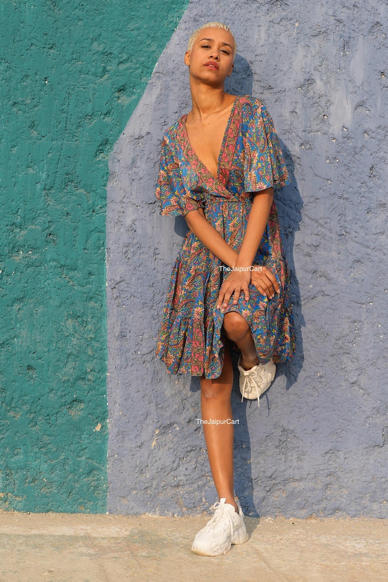 K&ouml;nnte beinhalten: Eine Frau tr&auml;gt ein blaues, rosa und orangefarbenes Kleid mit Blumenmuster, V-Ausschnitt und kurzen &Auml;rmeln. Das Kleid hat einen R&uuml;schensaum und wird mit wei&szlig;en Turnschuhen kombiniert. Der Hintergrund ist eine strukturierte Wand mit blauer und t&uuml;rkisfarbener Farbe.
